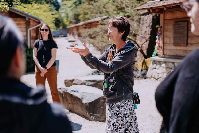 A person gesturing while speaking to a group of people outdoors, with traditional wooden buildings and a stone wall in the background. 1