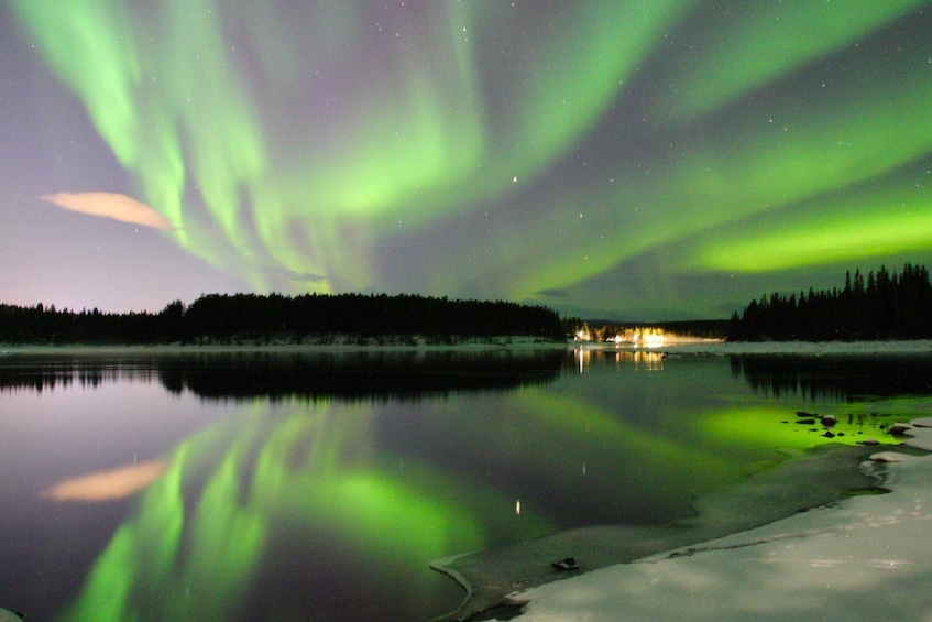 The Northern Lights over a calm lake with a forested shoreline and a distant lit building. 3