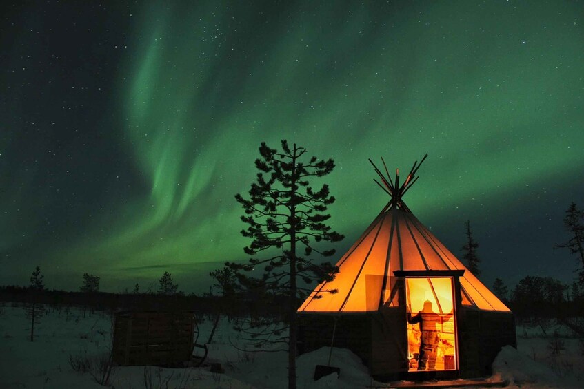 A snow-covered landscape with a teepee illuminated by a warm light, under a night sky with the Northern Lights. 1