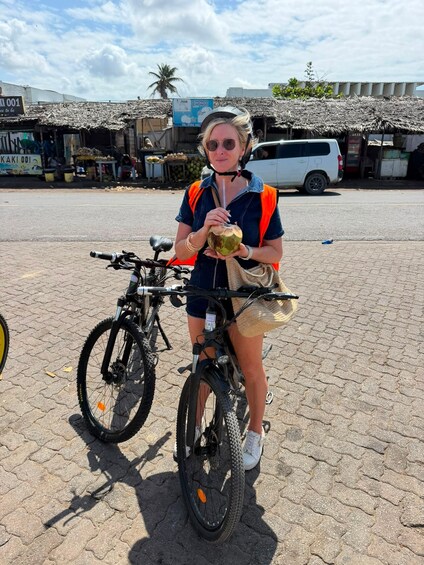 A person on a bicycle with a basket, holding a coconut, in an outdoor market area with buildings and a van in the background. 4