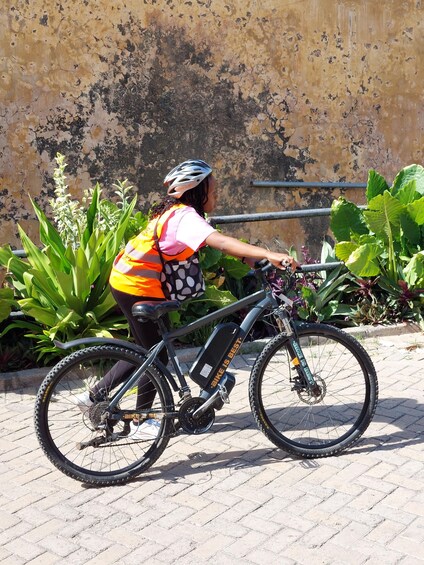 A person riding a bicycle with a safety vest and helmet, parked next to a wall with plants. 5