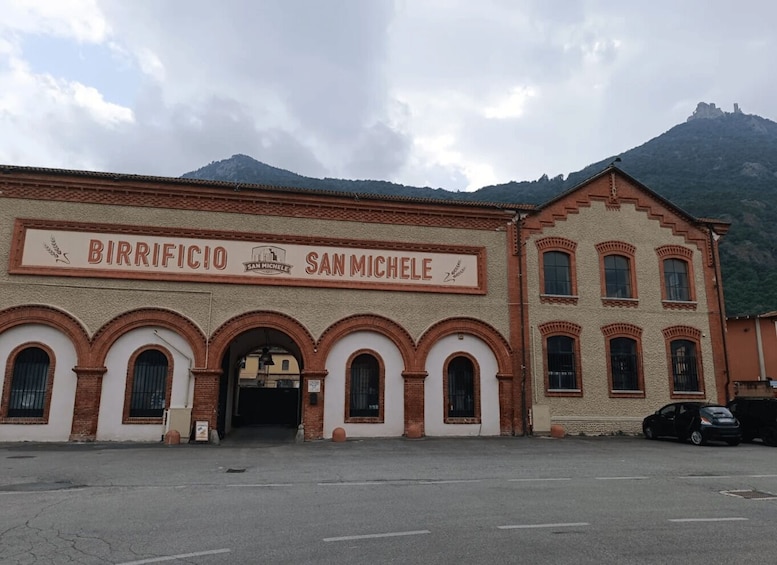 A building with 'BIRRIFICIO SAN MICHELE' signage, arched windows, and a mountainous backdrop. 5