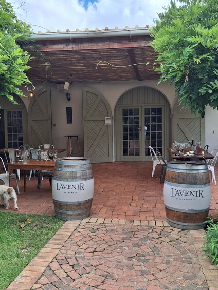 A rustic outdoor dining area with brick flooring, two large L'avenir beer barrels, a wooden table set with glasses, and a dog nearby. 4