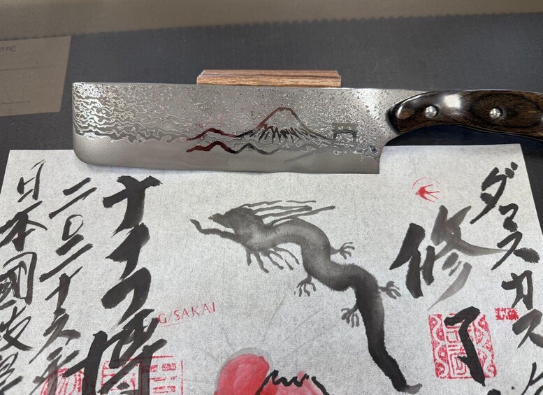 A traditional Japanese sword with a wooden handle, a metal blade with a mountain design, and a piece of paper with calligraphy and a red seal. 2