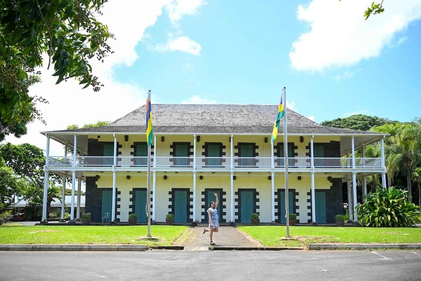 A colonial-style building with a balcony, two flags, and a person standing in front. 4