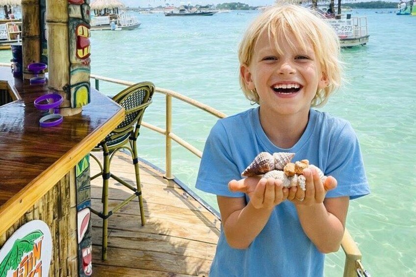 Eine Holzterrasse mit Geländer, ein Kind das Muscheln hält, und ein Gewässer im Hintergrund mit Booten. 1