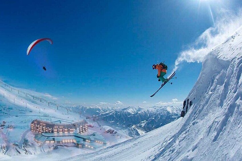 Een skiër maakt een sprong op een besneeuwde helling, met een skigebied op de achtergrond. 1