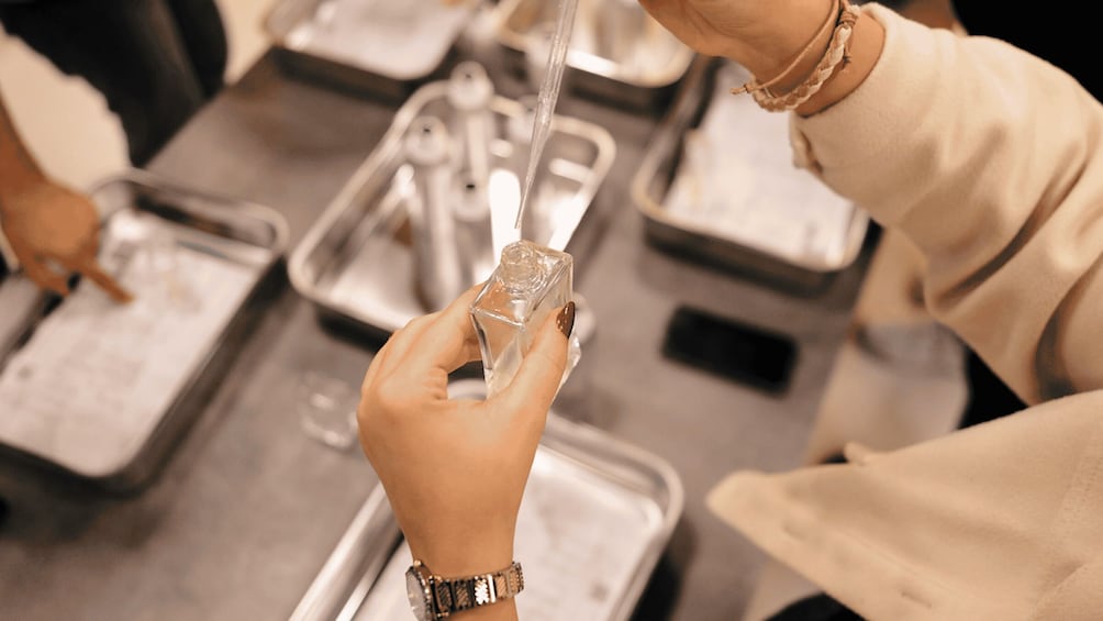 A person holding a perfume bottle, wearing a watch and bracelet, at a table with trays. 1