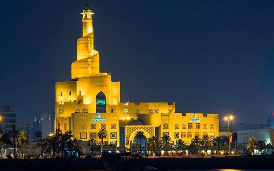 A large, illuminated building with a spiral minaret, surrounded by palm trees and other structures. 5
