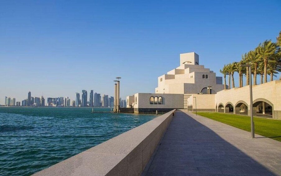 A waterfront promenade with a modern building, palm trees, and a city skyline in the background. 2