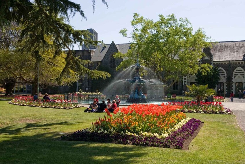 A park with a fountain, colorful flower beds, and people sitting on the grass. 3
