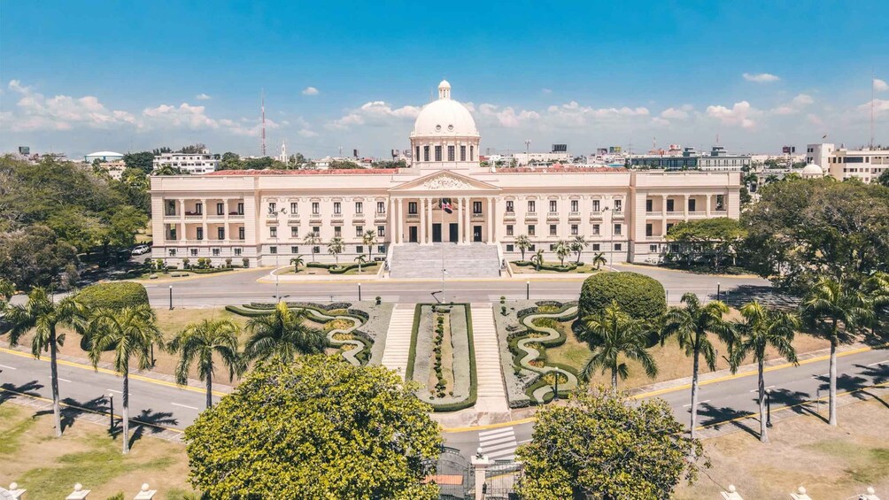 A large, white, neoclassical building with a central dome and symmetrical facade, surrounded by manicured gardens and palm trees. 3