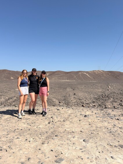 Trois personnes se tiennent sur un terrain rocheux, sous un ciel dégagé, avec des collines qui se dessinent à l’horizon. 2