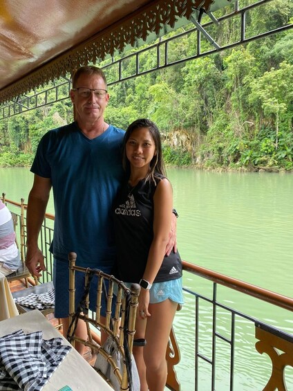 A man and a woman standing by a river with a wooden railing and greenery in the background. 3