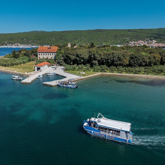 A boat named 'Kosljun Tour' on a clear day, docked at a pier with a building and greenery in the background. 1