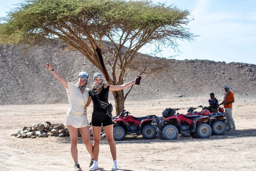 Two people taking a selfie with a tree and ATVs in a dry, rocky landscape. 4