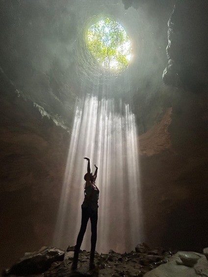 A person standing in a cave with a light source above, casting rays down. 1