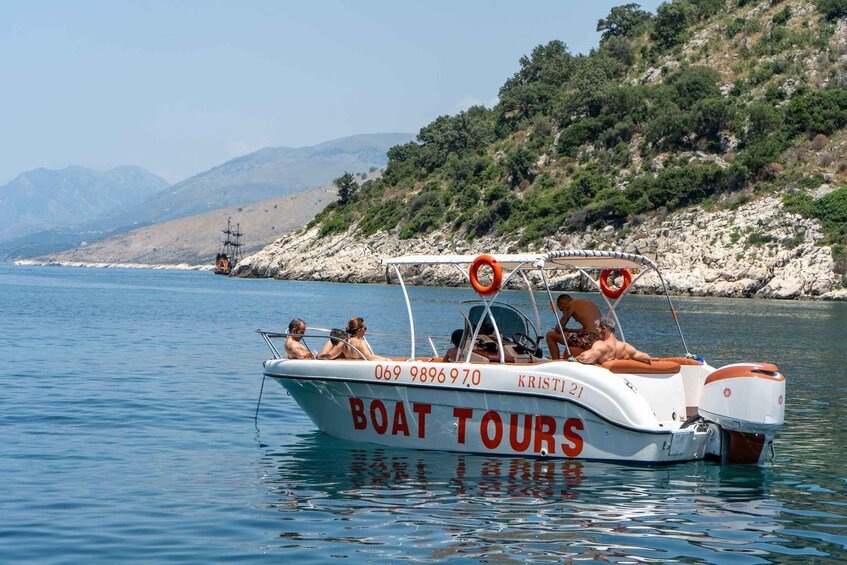 A boat tour named "Boat Tours" with the number 069 9896970 and "Kristi 21" on its side, floating on calm waters with a rocky coastline and hills in the background. 5