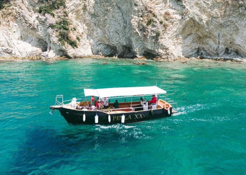 A boat named 'PELLA XX' with a white canopy, sailing on turquoise water near a rocky coastline. 3