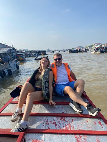 Two people sitting on a red and white dock in a waterway with boats and buildings in the background. 5