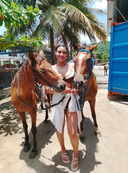 A person standing between two horses in a fenced area with palm trees and a blue structure in the background. 1