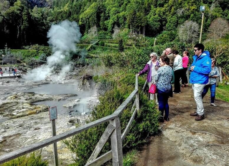 A group of people standing on a wooden boardwalk overlooking a geothermal area with steam rising from the ground. 1