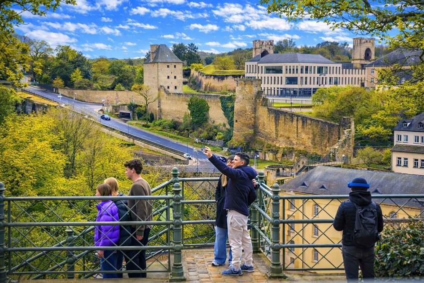 A group of people taking a selfie on a bridge overlooking a historic castle and modern buildings. 1