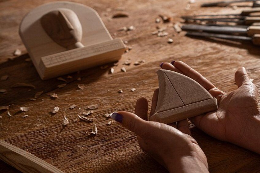 Hands holding a wooden object, a wooden stamp, and a wooden block on a wooden surface with wood shavings. 4