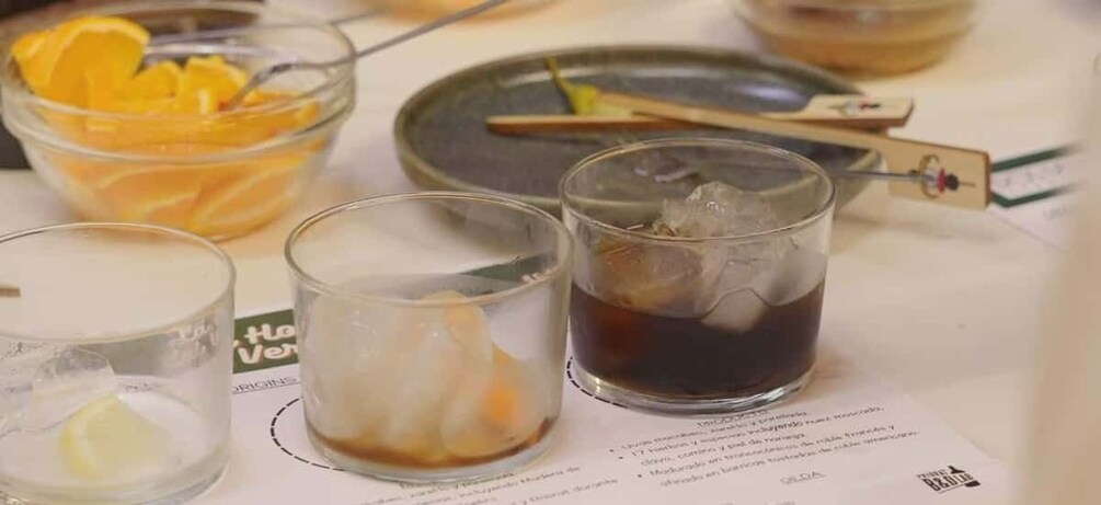 Three glasses of beverages on a table with a bowl of orange slices and a plate with a wooden spoon. 5