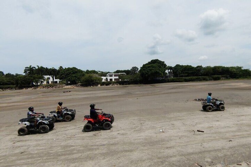 Four people riding atv's on a sandy road with buildings and trees in the background. 1