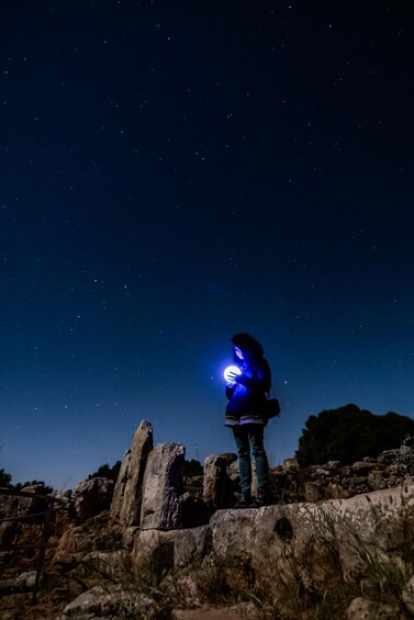 A person standing on rocky terrain under a starry night sky, holding a flashlight. 4