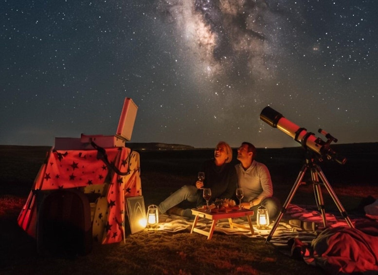 Two people stargazing with a telescope and a picnic setup under a starry sky. 1