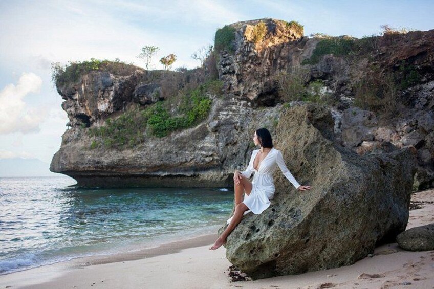 A person sitting on a large rock by the sea, with cliffs and vegetation in the background. 5