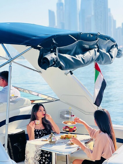Two people dining on a boat with a table set for tea and snacks, a flag, and a city skyline in the background. 3