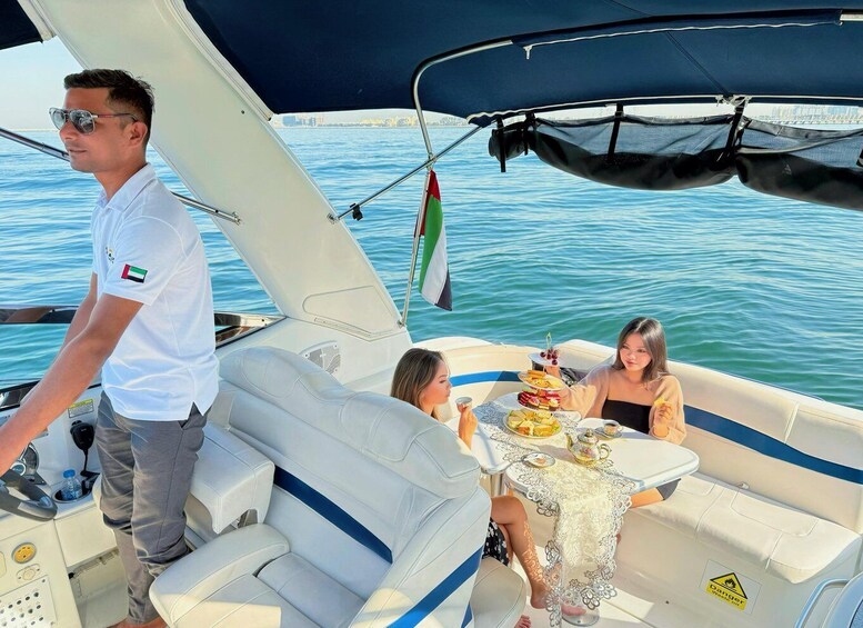A boat with two passengers and a man steering. There's a table with food and drinks, and a flag is visible. 2