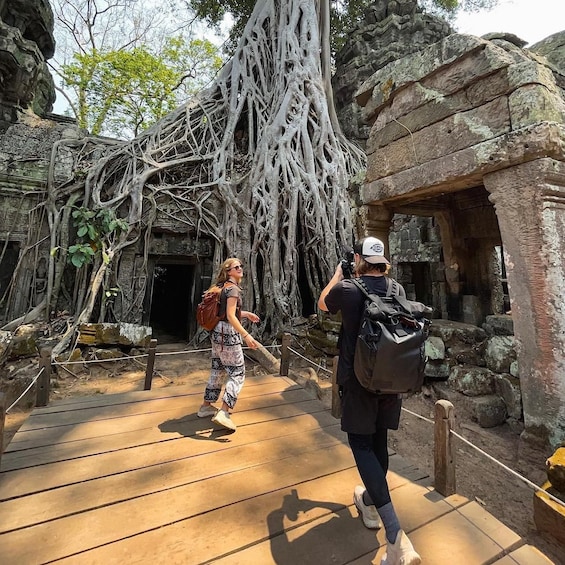 A person taking a photo of a tree with extensive roots in front of a stone structure. 1
