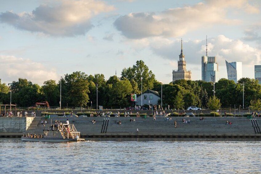 A riverside park with people relaxing and walking, a historic tower, and modern buildings in the background. 2