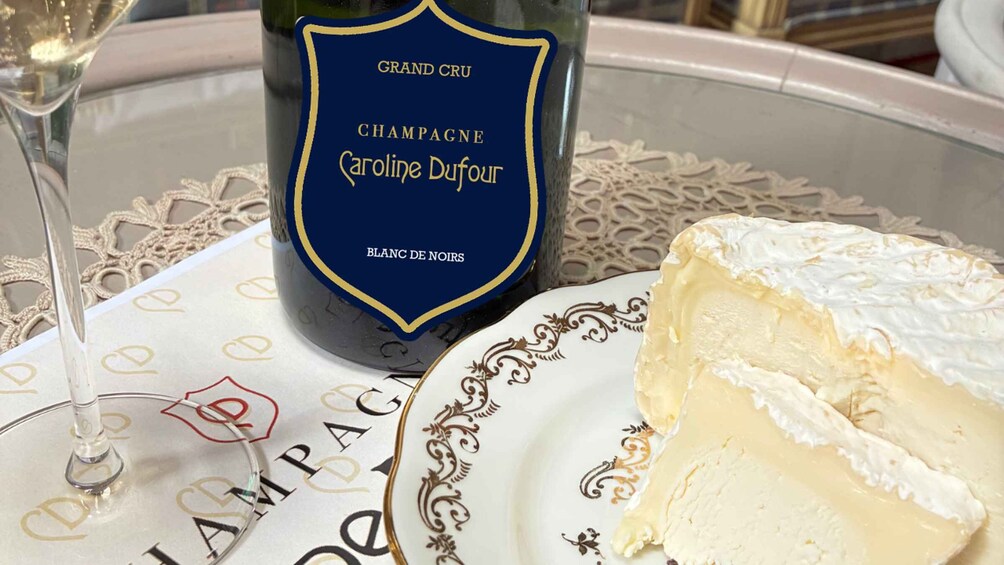 A bottle of champagne, a glass of champagne, and a piece of cheese on a decorative plate. 1
