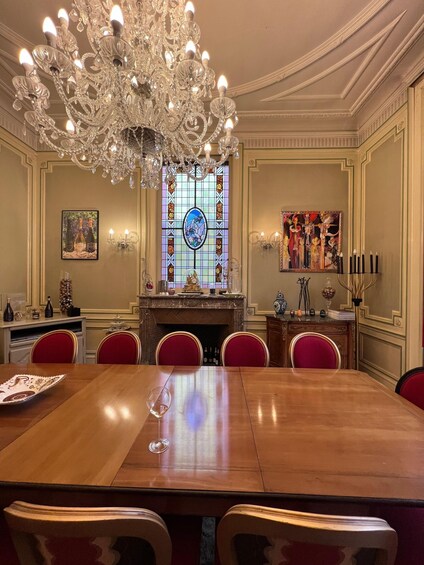 A formal dining room with a large wooden table, red upholstered chairs, a crystal chandelier, a stained glass window, and a fireplace. 2