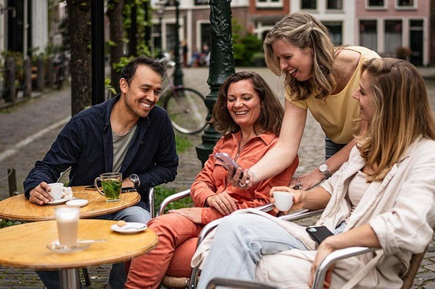 A group of people sitting at a round wooden table outdoors, with a bicycle and buildings in the background. 2
