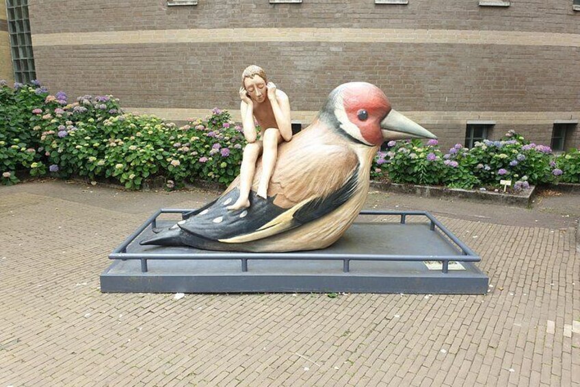 A sculpture of a bird with a human figure sitting on it, set on a platform in an outdoor area with brick walls and flower beds in the background. 5