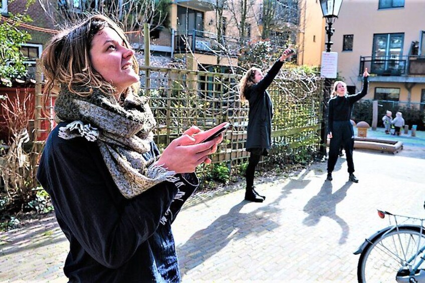 A person holding a smartphone, another person taking a photo, and a bicycle in an outdoor setting with buildings and a fence in the background. 3