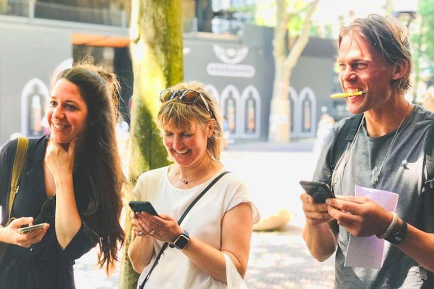 Three people standing outdoors, one holding a smartphone, another holding a drink, and the third looking at a smartphone. 1