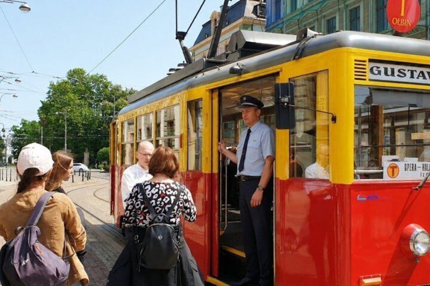 Eine Straßenbahn, auf der Fahrgäste einsteigen, ein Straßenbahnbetreiber und ein Schild mit der Aufschrift 'GUSTAV'. 1