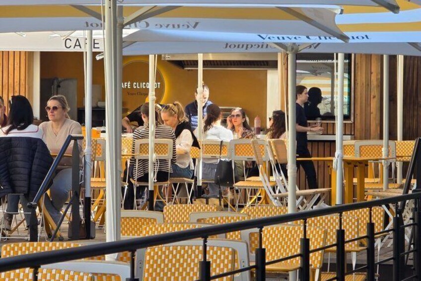 An outdoor café with yellow checkered chairs and tables, some people seated, and a few standing. 2