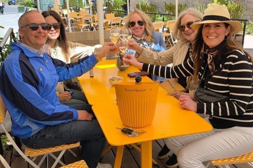 A group of people toasting with wine glasses at a yellow table with a bottle of Veuve Clicquot. 1