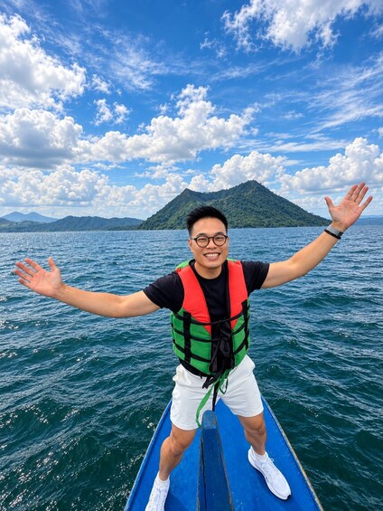 A person on a blue boat with arms outstretched, wearing a life jacket, white shorts, and white shoes, with a mountainous landscape and a blue sky with clouds in the background. 5