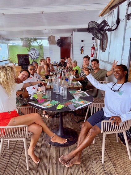 A group of people seated around a round table with drinks, in an outdoor setting with a ceiling fan and a fire extinguisher visible. 4
