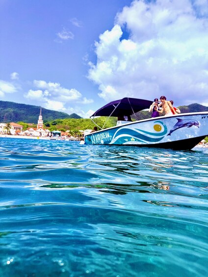 A boat with a dolphin design on its side, sailing on clear blue water with a church and mountains in the background. 2