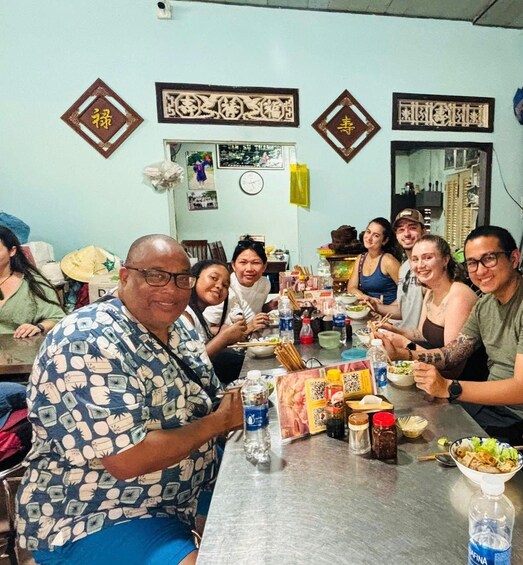 A group of people sitting around a table with food and drinks, in a room with decorative wall hangings. 2
